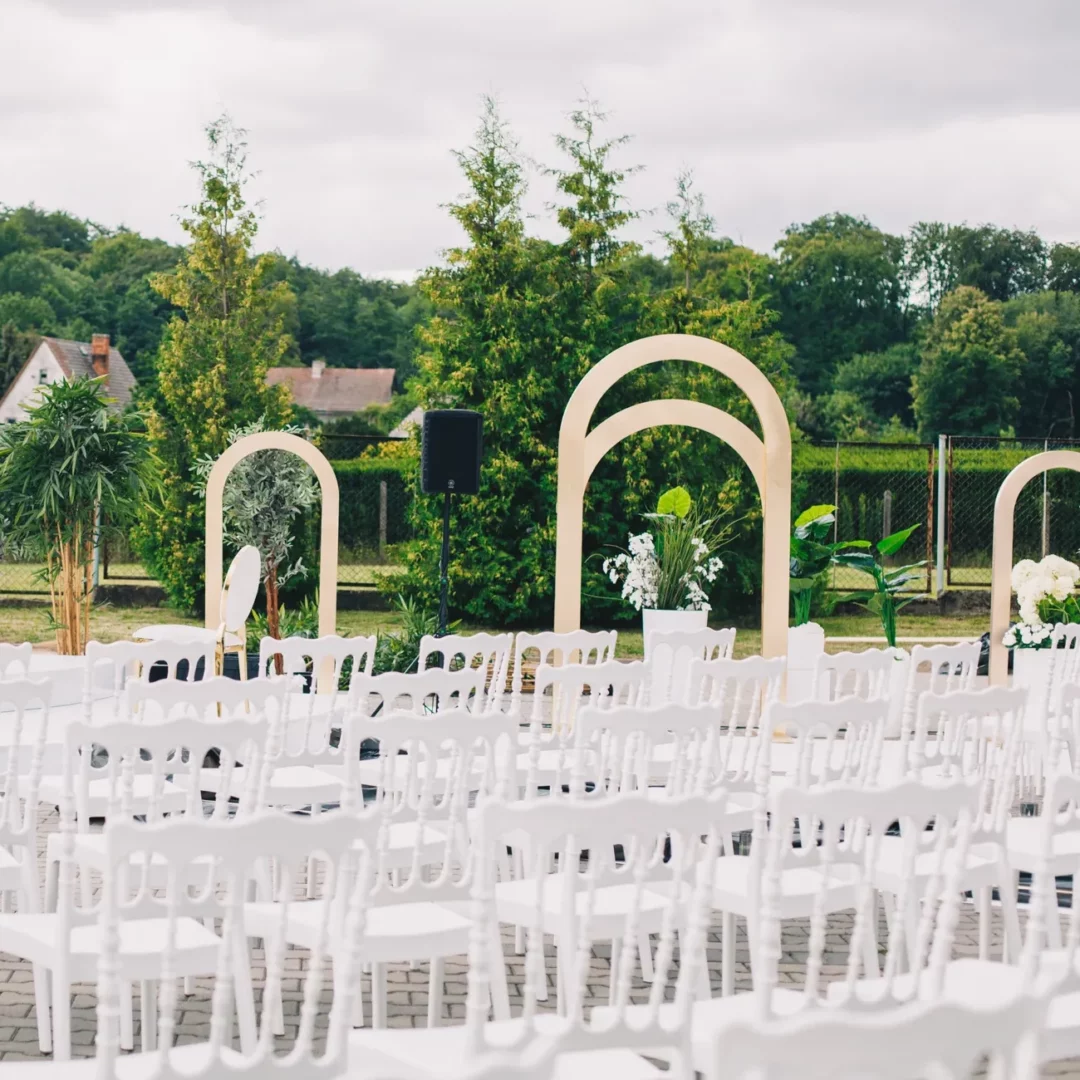 Plenerowa uroczystość w otoczeniu zieleni podkreślona przez białe krzesła Napoleon i drewniane łuki ślubne to propozycja od RENT4WEDDING na wyjątkowe śluby i eventy we Wrocławiu i okolicach. Minimalistyczne, ale zarazem stylowe dekoracje komponują się z naturą, tworząc idealne tło dla wymarzonych chwil - rent4events.pl wypożycz wrażenia, rent4wedding.pl zachowaj wspomnienia.