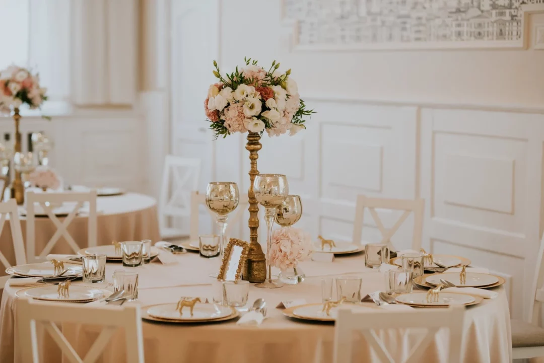 Aranżacja stołów na eleganckim weselu z centralnym złotym świecznikiem/kwietnikiem z pięknymi różowymi kwiatami, złotymi talerzami i sztućcami, prezentowane przez rent4events.pl. Jasna i przestronna sala z białymi ścianami oraz beżowymi obrusami idealnie komplementuje złote i różowe akcenty, tworząc ekskluzywną atmosferę. Numer stołu "4" w złotej ramce dodaje personalizacji i stylu - rent4events.pl wypożycz wrażenia, rent4wedding.pl zachowaj wspomnienia.