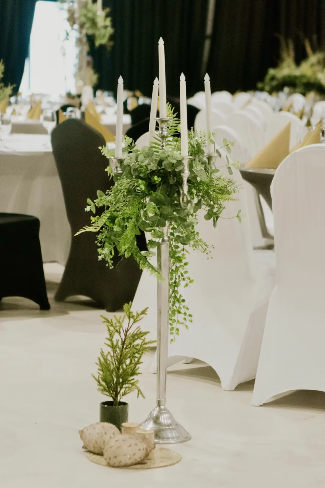 Pęcioramienny srebrny kandelabrem 100 cm, otoczonym wiankiem zielonych roślin, z rent4events.pl. Elegancka kompozycja światła i natury na tle eleganckich stołów w biało-czarnej kolorystyce, podkreśla styl klasyczny i prestiżowy. Idealna dekoracja do stworzenia niezapomnianego klimatu na weselach i uroczystych bankietach - rent4events.pl wypożycz wrażenia, rent4wedding.pl zachowaj wspomnienia. Fot. @karolinakiec_fotografia .