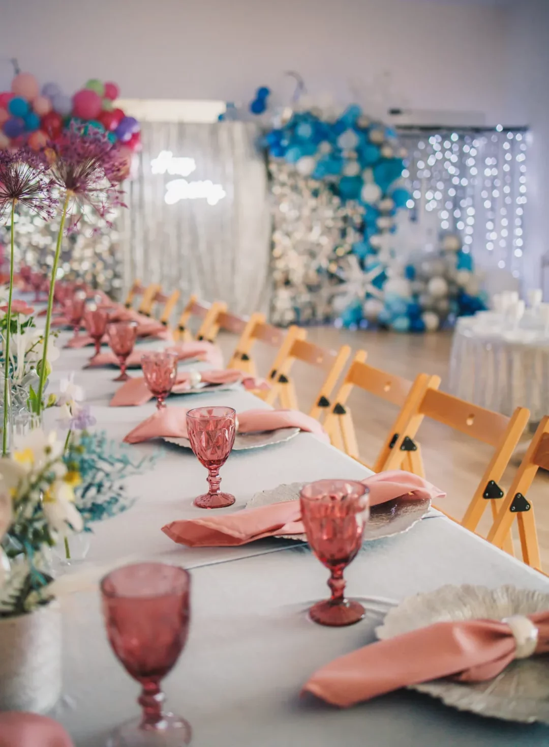 Elegancka aranżacja stołu rent4events.pl na wydarzenie we Wrocławiu z różowymi szklanymi kieliszkami i serwetkami, ułożonymi na srebrnych podkładkach. Tło zdobią kolorowe balony i błyszczące zasłony, tworząc wspaniałą scenografię idealną na wesele lub event, które rent4weddings.pl przekształca w trwałe wspomnienia - rent4events.pl wypożycz wrażenia, rent4wedding.pl zachowaj wspomnienia. Fot. @waterlily_photo.