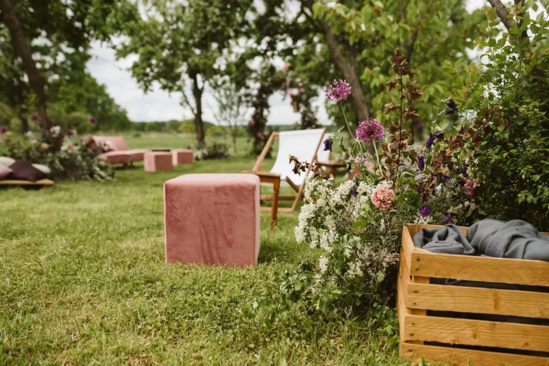 W malowniczym otoczeniu zielonego trawnika, elegancka różowa Pufa Kubik w pastelowym odcieniu oraz leżak z drewnianą ramą i białym płóciennym siedziskiem tworzą przytulne miejsce do relaksu. Obok znajduje się kompozycja kwiatowa z fioletowymi, różowymi i białymi kwiatami oraz drewniana skrzynka z szarym materiałem. W tle widoczna jest różowa sofa z poduszkami, umieszczona wśród zielonych drzew i krzewów, które dodają naturalnego uroku i prywatności. Ta scenografia to doskonały wybór na ślub, wesele czy każdy event, który wymaga eleganckiej i stylowej aranżacji. Idealna wypożyczalnia dekoracji we Wrocławiu - rent4events.pl wypożycz wrażenia, rent4weddings.pl zachowaj wspomnienia. Fot. @my.favourite.human .