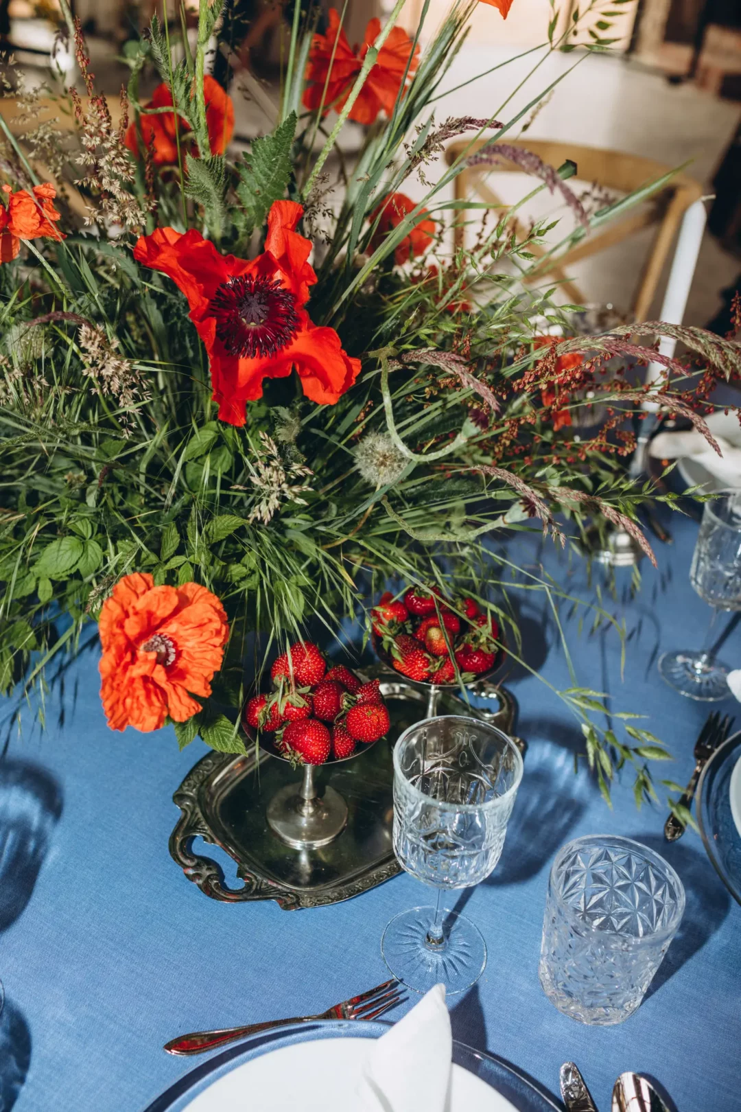 Zbliżenie na elegancką aranżację stołu weselnego, ukazujące bukiet czerwonych maków i zieleni na niebieskim obrusie Dusty Blue. Bukiet jest uzupełniony srebrną tacą z dekoracyjnymi naczyniami wypełnionymi truskawkami, dodającymi kolorowego akcentu. Obok znajdują się kryształowe szklanki i kieliszki oraz biała zastawa stołowa z elegancko złożonymi serwetkami. Drewniane krzesła z jasnym tapicerowanym siedziskiem harmonizują z dekoracjami stołu, tworząc spójną i stylową kompozycję. Scenografia ta jest idealna na ślub i wesele - rent4events.pl wypożycz wrażenia, rent4weddings.pl zachowaj wspomnienia. Fot. Norma Ilnicka @luckyinlovepl.