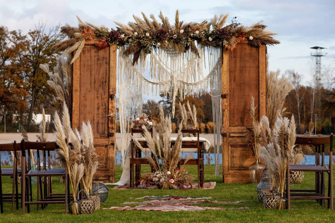 Stylowa aranżacja miejsca ceremonii ślubnej w plenerze, w rustykalnym stylu z centralnym punktem w postaci dwóch starych drewnianych drzwi, otoczonych licznymi dekoracjami z traw pampasowych. Wiszące girlandy z suszonych kwiatów i traw dodają romantycznego i eleganckiego charakteru. Po obu stronach przejścia ustawione są drewniane krzesła, a za drzwiami znajduje się mały drewniany stół z kwiatową dekoracją. Przestrzeń przed drzwiami zdobią kolorowe dywany, wprowadzając element stylu boho i przytulności. Tło to zielony krajobraz z drzewami i jasnym niebem, sugerujące wczesną jesień. Ta scenografia to doskonały wybór na ślub, wesele czy każdy event, który wymaga eleganckiej i stylowej aranżacji - rent4events.pl wypożycz wrażenia, rent4weddings.pl zachowaj wspomnienia. Fot. @masterfilmwroclaw.