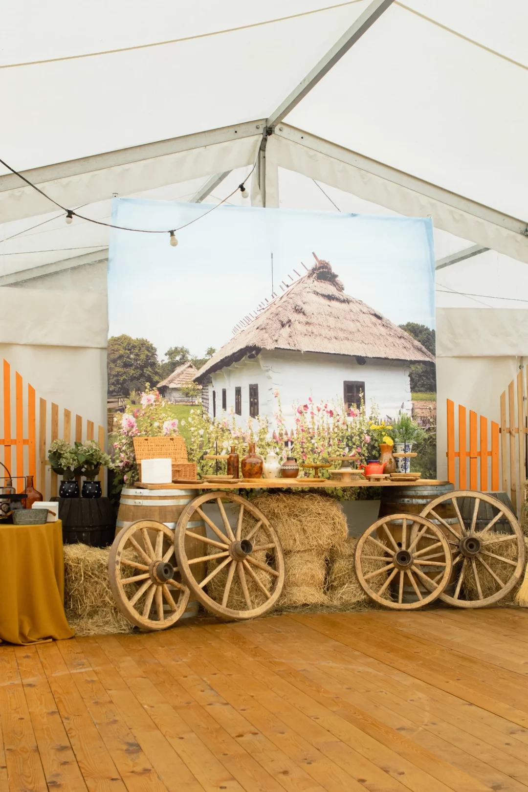 Horyzont Krajobraz Wieś Chata w sielskiej aranżacji. Rustykalna scenografia eventowa przedstawiająca tło fotograficzne z wiejskim domkiem z białymi ścianami i strzechą, otoczone drewnianymi kołami od wozu, beczkami oraz stołem udekorowanym słomą, naczyniami, butelkami i kwiatami. Całość zaaranżowana w namiocie z białym dachem i drewnianą podłogą, idealna na farmerskie wesele, ślub czy inne eventy w sielskiej atmosferze. Wypożyczalnia dekoracji we Wrocławiu oferująca wyjątkowe scenografie na eventy - rent4events.pl, zapewnia wrażenia, a rent4weddings.pl pomoże zachować wspomnienia. Fot. @karolinakiec_fotografia.