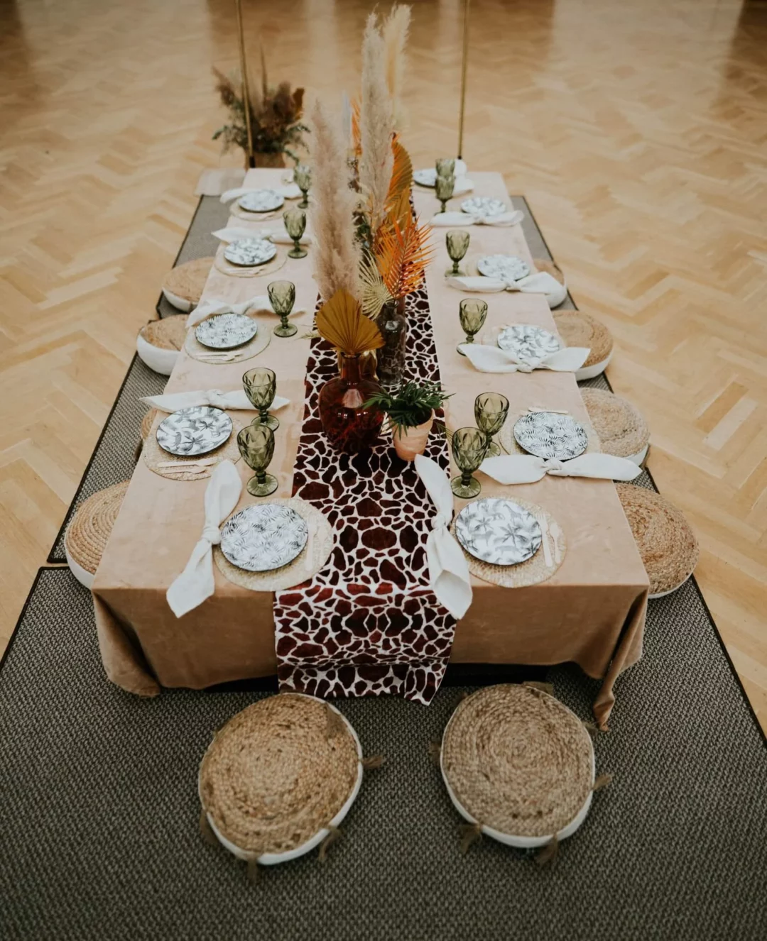 Stół w stylu Safari Boho, udekorowany bieżnikiem z motywem skóry żyrafy, suszonymi roślinami oraz elegancką zastawą stołową. Plecione Siedziska Boho z juty i bawełnianego obszycia w stylu boho leżą dookoła stołu, a zielone kieliszki i talerze z roślinnymi wzorami podkreślają naturalny, egzotyczny charakter dekoracji. Aranżacja doskonała na eventy, śluby oraz wesela organizowane we Wrocławiu i okolicach. Wypożyczalnia rent4events.pl oferuje szeroki wybór dekoracji, łączących styl safari z boho, tworząc oryginalne i eleganckie scenografie - rent4events.pl wypożycz wrażenia, rent4weddings.pl zachowaj wspomnienia